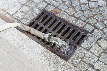 Firefighter hose draining water into metal street sewer grate. Emergency equipment in use during...