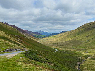 Scenic valley view with winding road between rolling green hills under a cloudy sky.