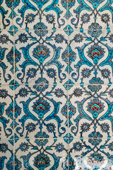Traditional Islamic ceramic tile wall decorated with turquoise and blue floral arabesque motifs in repeating patterns, ornamental detail from Middle Eastern architecture.