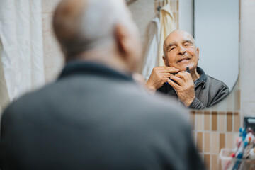 A senior man using an electric razor to shave while smiling and checking his reflection in the...