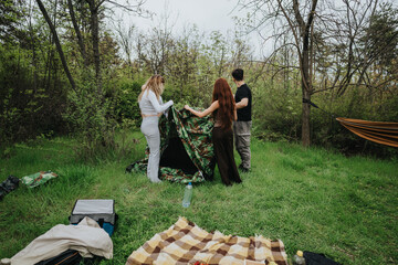 Friends enjoying camping as they assemble a tent surrounded by lush green nature. The scene conveys teamwork, outdoor leisure, and a serene connection with the natural environment in a wooded area.