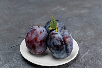 Three ripe blue plums in white saucer on grey background, fresh seasonal fruits