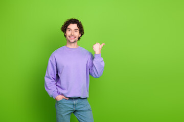 Smiling young man in purple sweatshirt gesturing with finger on vibrant green wall background,...