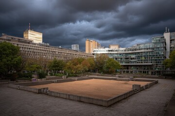 Fototapeta premium Empty square courtyard surrounded by modern buildings under a dramatic sky.