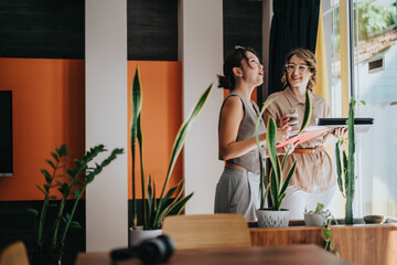 Two women are enjoying a casual interaction near growing plants in a bright, colorful interior. The...