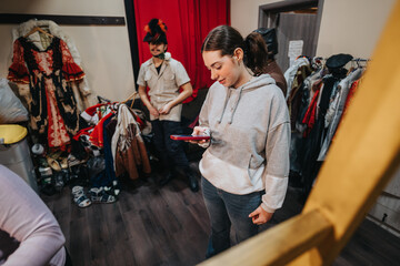 Two young individuals in a theater costume area, surrounded by colorful outfits, set in a creative and lively setting, preparing for a performance.