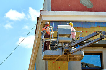 Two construction workers in crane bucket insulating and plastering exterior wall, facade renovation...