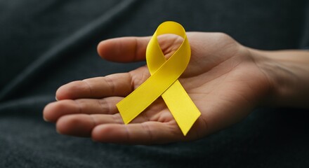 A hand holds a yellow ribbon in a bow shape The background is a dark fabric
