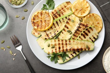 Delicious grilled courgette slices with pumpkin seeds, lemon and parsley served on grey textured table, flat lay
