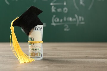 Scholarship. Rolled dollar banknotes with graduate hat on wooden table against green chalkboard,...