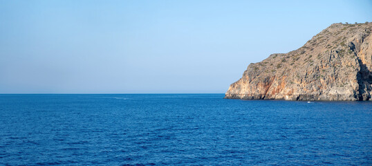 Fototapeta premium A rugged rocky cliff rises above the calm deep blue sea along the Greek coastline, captured within clear daylight featuring the serene horizon and soft natural tones
