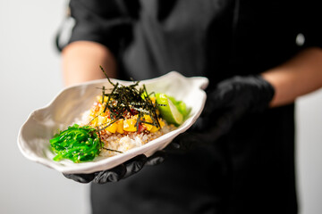 Gourmet poke bowl with rice, mango, tuna, avocado, seaweed salad, and nori in a white ceramic dish, held by gloved hands. Vibrant, fresh, and artistically plated