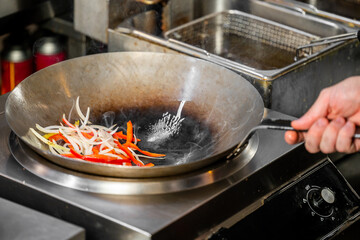 Chef stir-frying sliced bell peppers and onions in a hot wok over a gas burner in a professional kitchen. Steam rises, highlighting dynamic cooking in a commercial setting