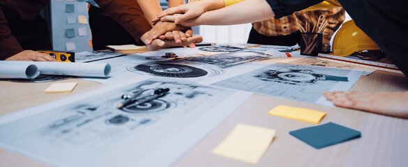 A diverse group engages in collaboration, placing hands together over architectural plans on a modern table, showcasing teamwork and creative problem-solving in the office environment. SACTR