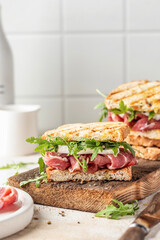 Grilled ham, mozzarella and arugula sandwich on wooden cutting board on white tile kitchen background close up. Delicious breakfast or snack