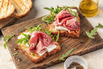 Grilled toasts with ham, cheese and arugula on wooden cutting board on light textured background close up. Delicious breakfast or snack