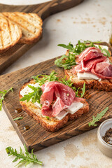 Grilled toasts with ham, cheese and arugula on wooden cutting board on light textured background close up. Delicious breakfast or snack