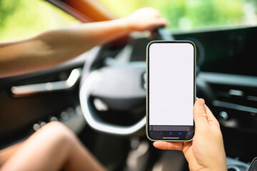 Close-up of female hand holding smartphone with white mockup on screen, background of car steering...