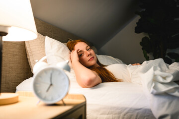 Young redhead woman on bedroom at home with clock close to