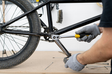 Fototapeta premium A person in gloves is tightening a bicycle crank on a bike frame using a ratchet wrench in a well-equipped workshop with tools in the background.