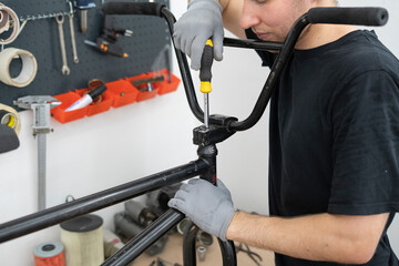 A person is carefully installing new bicycle handlebars in a workshop, using a screwdriver while wearing gloves. The bike rests on a stand for easy access.