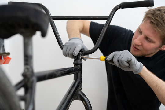 A person is carefully installing new bicycle handlebars in a workshop, using a screwdriver while wearing gloves. The bike rests on a stand for easy access.