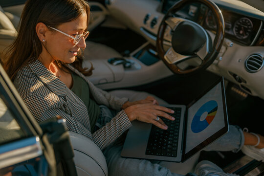 On a bright sunny day, a dedicated and focused businesswoman is working on her laptop inside a luxurious car