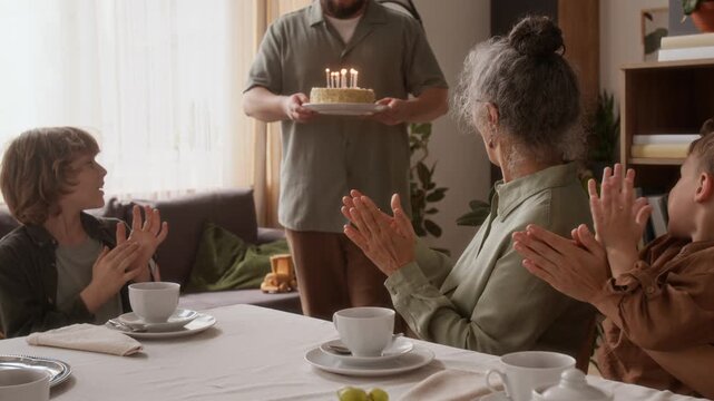 Multigenerational family clapping hands when man bringing birthday cake and placing it on festive table, senior woman blowing out candles and taking present from pre-teen grandson during home party