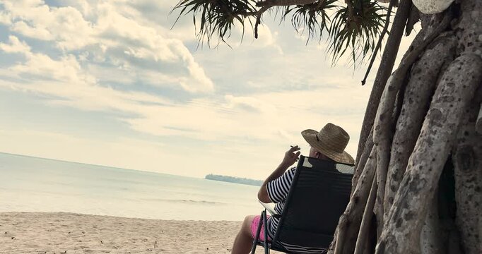 A man sits on a beach chair and smokes a cigarette on a sandy beach by the sea in a tropical country. Smoking weed on the beach, smoking cannabis by the sea. 