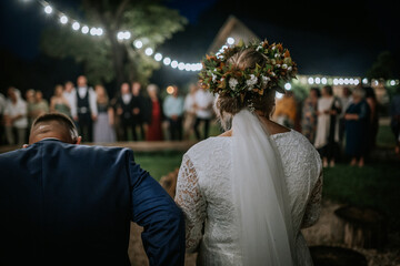 A bride and groom seen from behind during a nighttime outdoor wedding. The bride wears a flower...