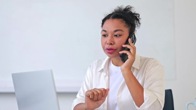 Confident professional businesswoman on phone call in modern office. Dressed in white shirt, woman works on laptop, demonstrating efficiency and focused communication. Bright, positive environment.