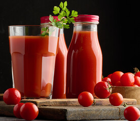 Full glass of tomato juice and a sprig of parsley behind a bottle of juice on a black table