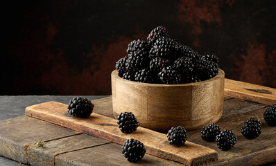 A wooden bowl filled with ripe blackberries on a board. A few berries are scattered around