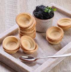 Close-up of black caviar in ceramic pot and mini tartlet placed on wooden board.