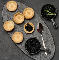 Black caviar in cast iron pans and tartlet on dark slate board. Empty tartlets and rosemary.