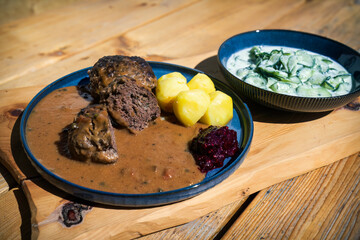 ready to eat -meatballs from chamois meat, potaoes, cranberry jam, brown sauce and cucumber salad, on a blue plate served on a wood board