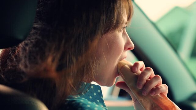 Beautiful woman eating tasty hot dog while sitting in car. Snack on road. Hungry girl eating hot dog sandwich from fast food in car. Woman satisfying hunger in car. Young woman having snack in car