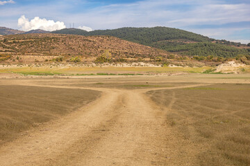 Dry countryside path leading through arid fields toward green hills under a blue sky
