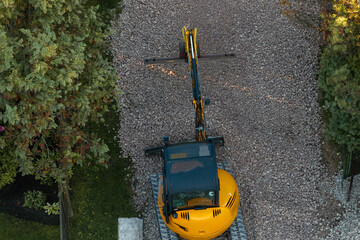 A yellow excavator sits stationary on a gravel pathway, viewed from above. Its arm is extended forward, surrounded by trees and bushes.