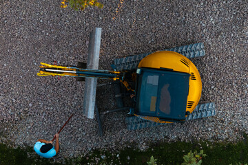 A yellow excavator with a black cabin holds a concrete beam on gravel. A person in a light blue shirt stands nearby, with vegetation at the frame's edge.