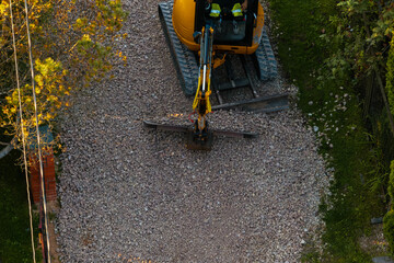A yellow excavator with an extended arm and flat attachment sits on gravel, surrounded by green vegetation and a red brick structure in sunlight.