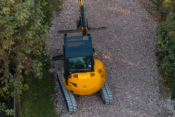 A yellow excavator with rubber tracks sits on a gravel pathway, viewed from above, with its arm extended forward and trees and bushes nearby.