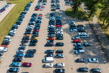 An elevated perspective of a parking lot with neatly arranged cars, various colors and models, streetlights, and trees lining the edges.