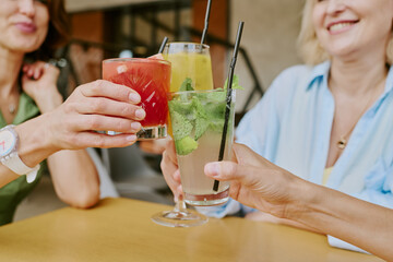 Fototapeta premium Three middle aged Caucasian women clinking glasses with colorful cocktails while smiling at table, hands and partial faces visible, celebrating friendship or special occasion