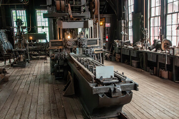 Inside a historic workshop, large vintage machinery stands prominently on wooden flooring. Sunlight streams through tall windows, illuminating the intricate tools and equipment used for craftsmanship.