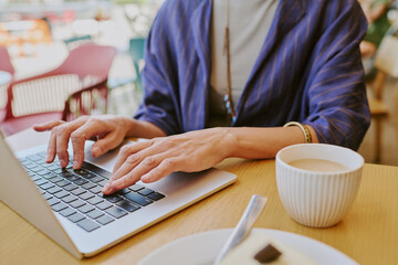 Caucasian young adult woman working on laptop at table, hands typing on keyboard with cup of coffee nearby, partial body visible, focusing on technology use in casual setting