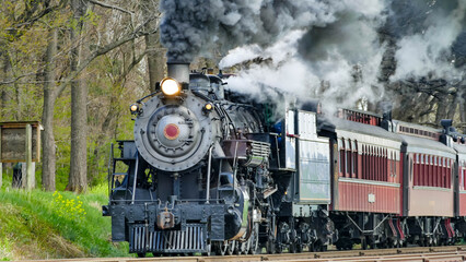 A historic steam locomotive engine pulls vintage passenger cars along tracks surrounded by lush green trees. Thick black smoke billows from the engine as it moves through the countryside.