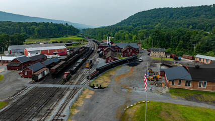 A vintage railroad station sits nestled among green mountains. Several trains are parked along the tracks. The area is quiet except for the distant sounds of nature.