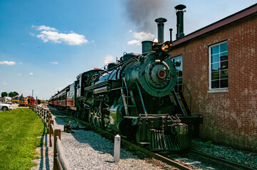 A classic steam locomotive stands proudly at a historic train station on a sunny summer afternoon. Passengers are visible, enjoying the nostalgic atmosphere while the train emits smoke.