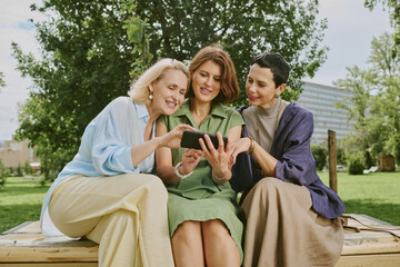 Three Caucasian middle aged women sitting outdoors on bench smiling and looking at smartphone...
